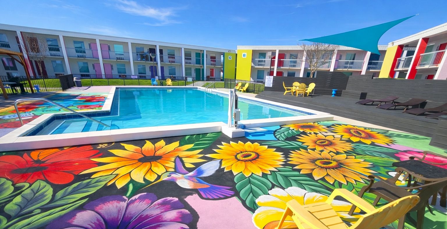 A colorful hotel pool area with a bright mural of oversized flowers in the foreground and lounge chairs; modern buildings surround the pool.