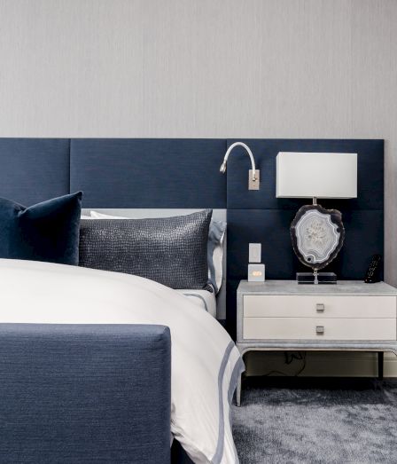 A modern hotel bedroom with a bed, blue headboard, and a nightstand holding a lamp, decorative geode, clock, and remote.