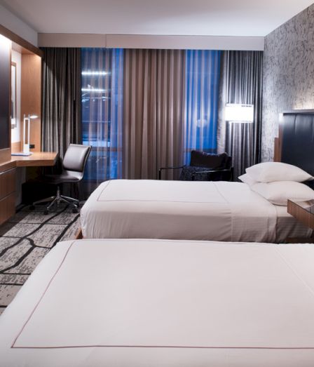A modern hotel room with twin beds, a desk chair, and curtains with lamps by the headboards.