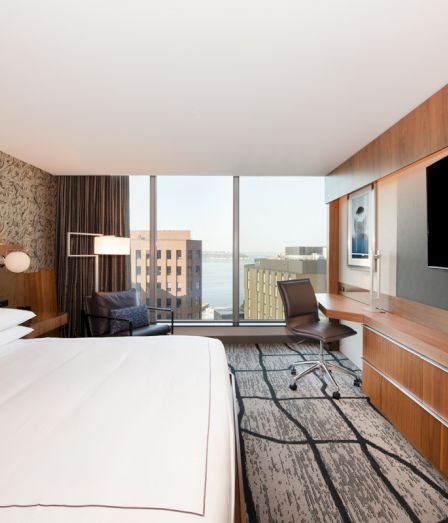 A modern hotel room with a bed, desk and chair, and city views through large windows, with sleek wood cabinetry.