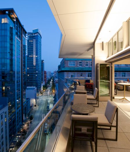 A modern rooftop terrace overlooks a city skyline, with outdoor seating along a bright glass-walled balcony.