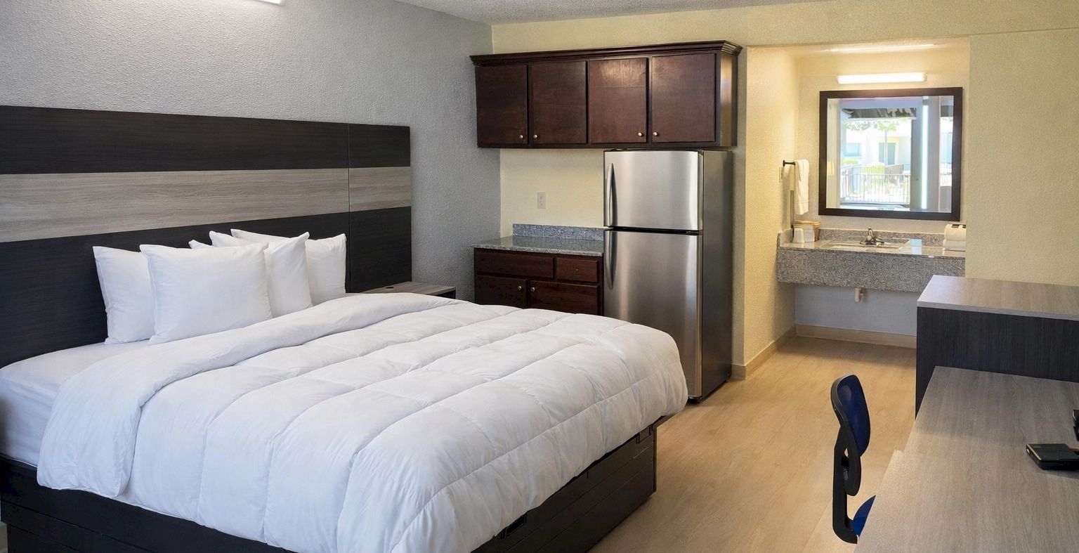 A modern hotel room with a large bed, white bedding, headboard, dark wood furniture, a kitchenette area with a fridge, and a desk facing a window.