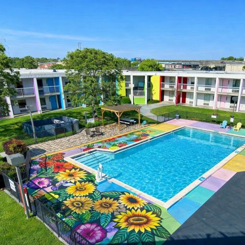 A colorful hotel courtyard with a rectangular turquoise pool, vibrant flower designs on the pool deck, around-the-pasin colorful buildings, green lawns, trees, and outdoor seating.