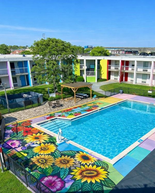 A colorful hotel courtyard with a rectangular turquoise pool, vibrant flower designs on the pool deck, around-the-pasin colorful buildings, green lawns, trees, and outdoor seating.