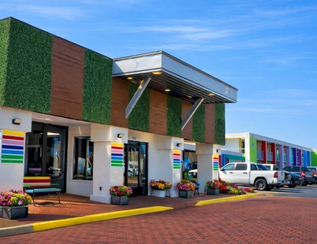 Colorful strip mall with green faux walls, striped awnings, potted plants, and parked cars along a brick-paved lot.