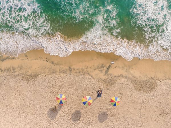 Three beach umbrellas and three people sitting on the sand by the waves, with footprints nearby and turquoise water washing ashore.