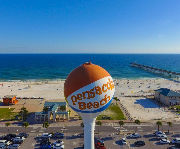 A crowded beach with umbrellas and sunbathers, a sign in the foreground by the shoreline, and blue sea under a clear sky, all on a sunny day.