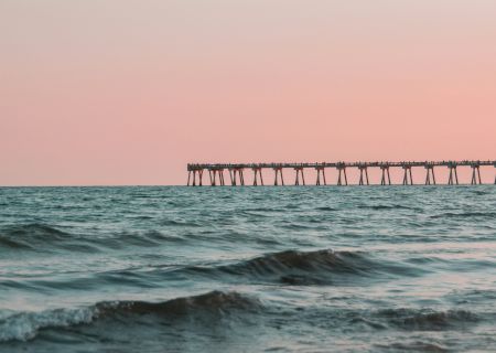 A long pier stretches out over choppy waves at sunset, silhouetted against a pale sky.