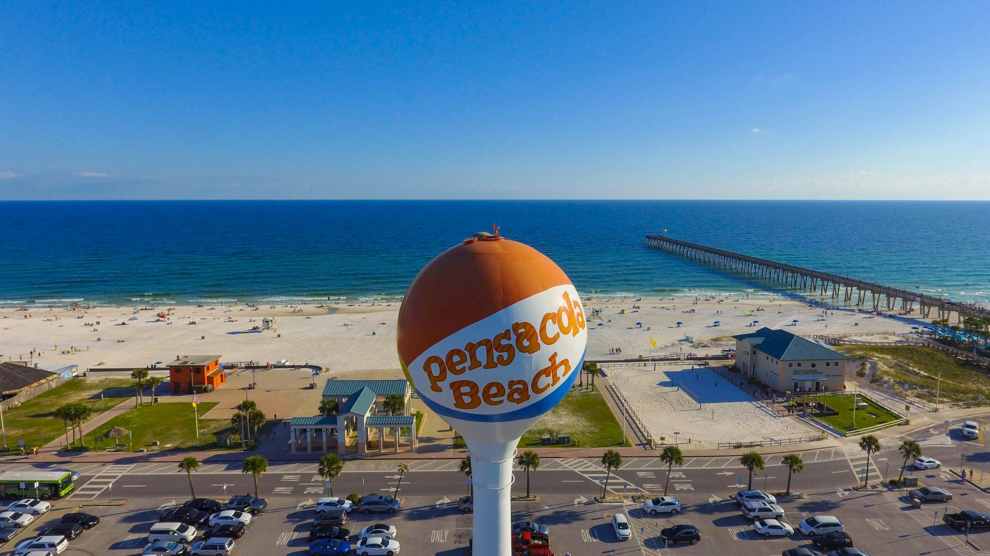 Aerial view of a sunny beach with a water tower labeled "Pensacola Beach," a parking lot, beach houses, and a long pier extending into the ocean, clear blue sky.