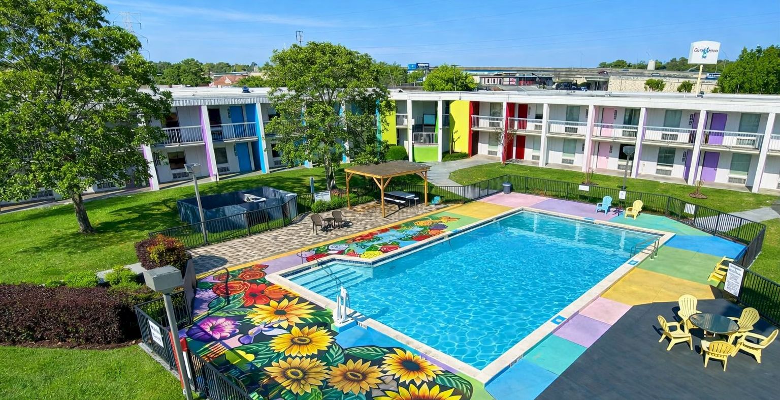 A colorful hotel courtyard with a bright rectangular pool, sun loungers, shade structures, and a cheerful building facade with rainbow accents.