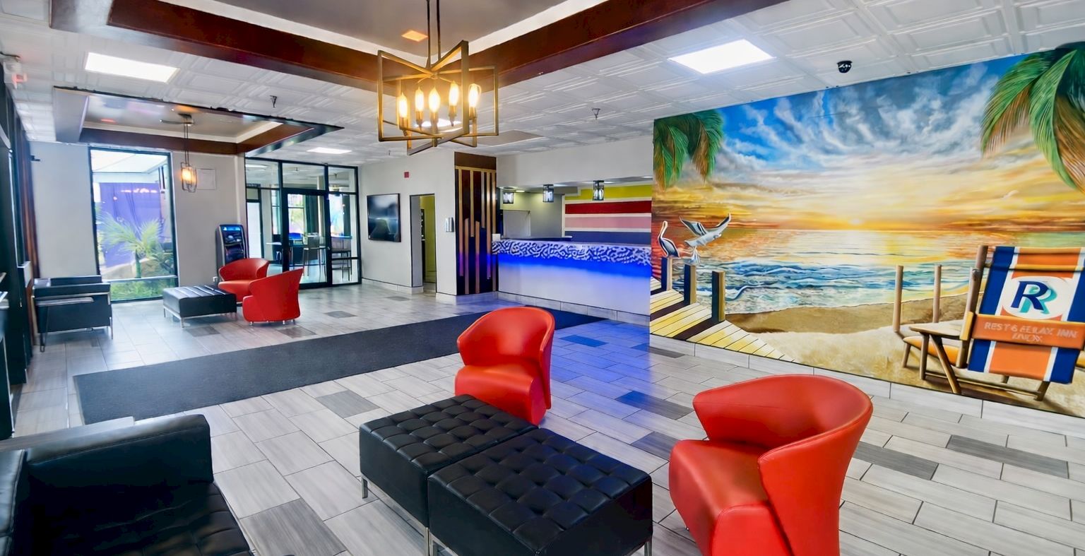 A modern hotel lobby with a beach mural, red accent chairs, black ottomans, white tiled floor, and a bright chic seating area across from a reception desk.