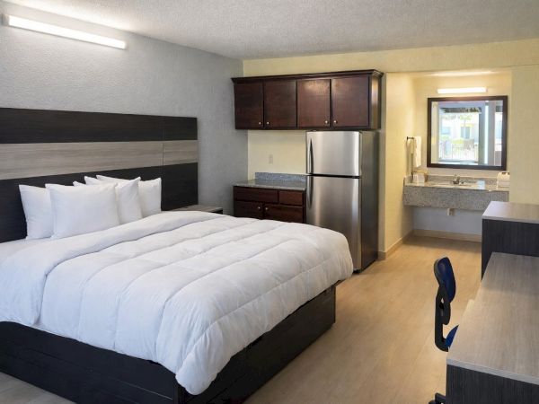 Modern hotel room: king bed with white linens, dark wood furniture, small kitchenette with fridge, desk, chair, and an adjoining sink area.