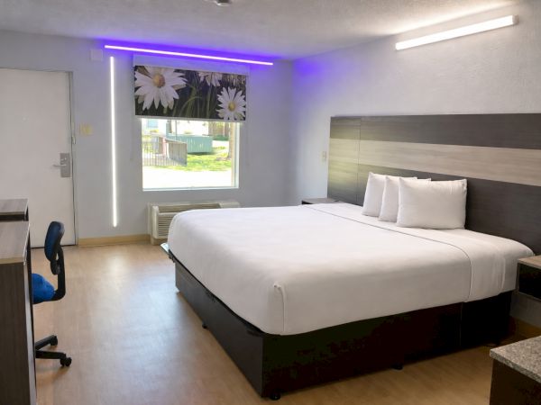 A clean hotel room with a large bed, minimalist dark wood furniture, a desk chair, window with blinds, and subtle purple LED lighting on the wall.