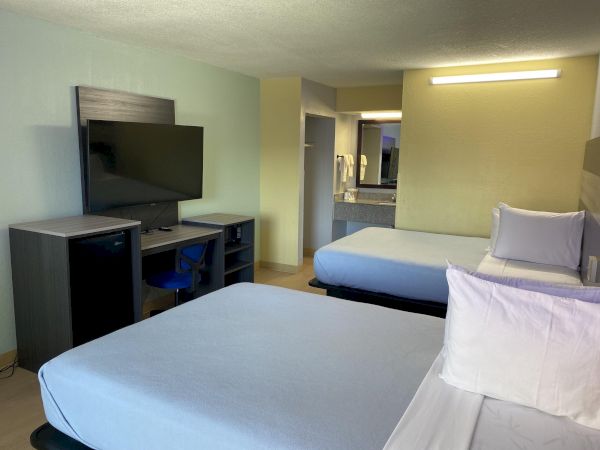 A hotel room with two queen beds, a mounted flat-screen TV on a dresser, a small desk area, and an open doorway leading to a hallway and kitchenette.