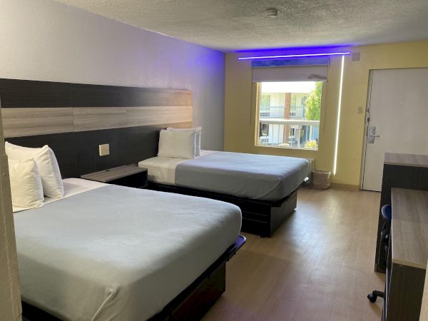 A hotel room with two beds, a window, desk, and a small nightstand; ceiling lights cast a blue glow near the window.