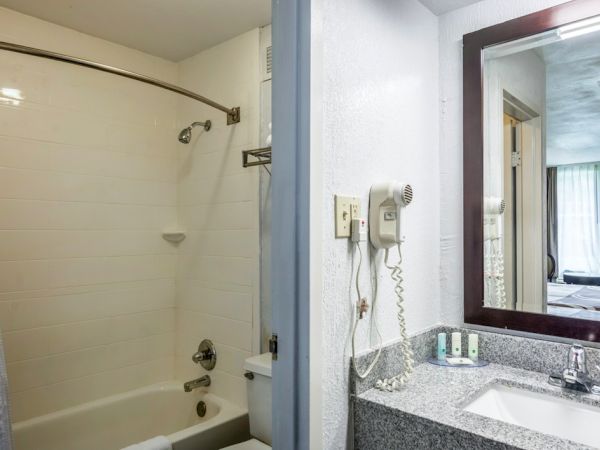 A small hotel bathroom with a tub/shower, curved shower rod, white tile walls, and a granite countertop with a sink, mirror, and wall-mounted phone, ending here.