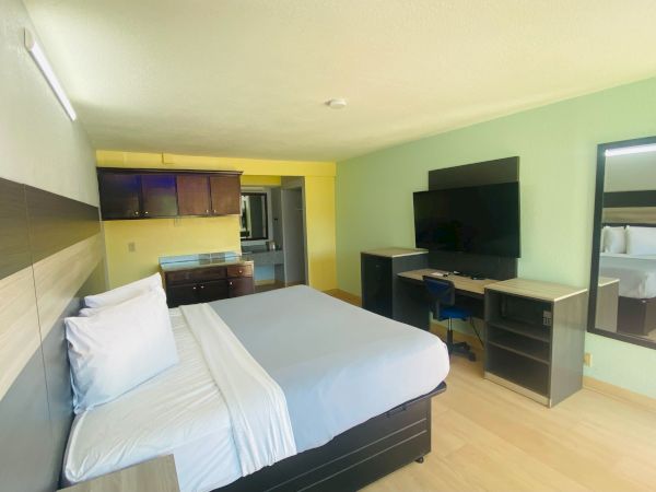 A modern hotel room with a king bed, wall-mounted TV, desk, mirror, and a small kitchenette in the background, bright and clean.