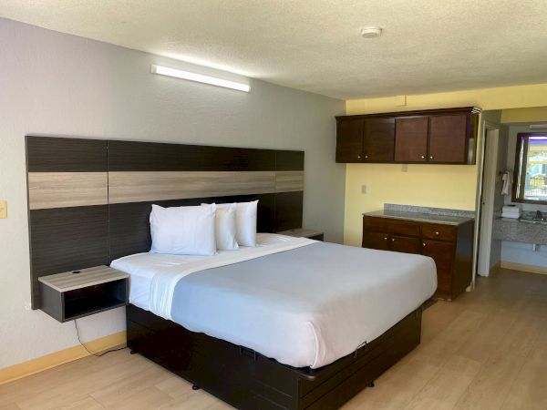 A hotel room with a large bed, dark wood headboard, built-in nightstands, and a small desk area; light wood flooring and a kitchenette in the back.