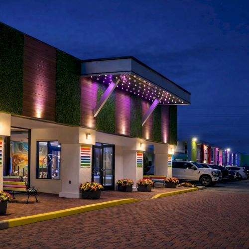 The image shows a row of colorful, two-story townhouses along a street at dusk, with warm lights glowing from the windows and a calm, clear sky.