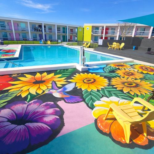 A sunny poolside scene with lounge chairs, colorful bean bags, umbrellas, and a bright hotel or resort building in the background, by the water.