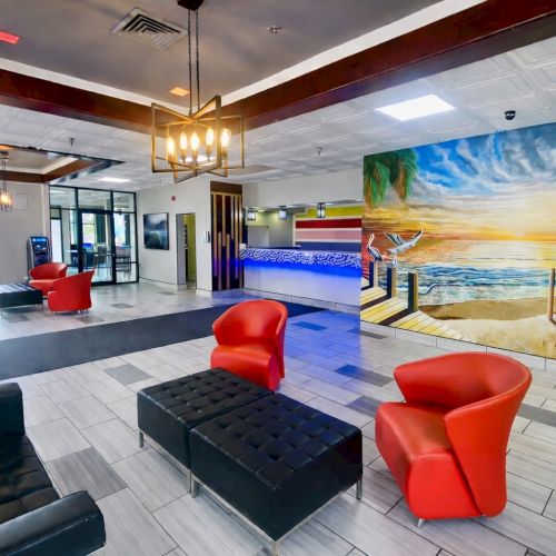 A modern hotel lobby with bright seating: red armchairs, black ottomans, light tiles, a tropical sunset mural, and a poolside vibe in the background.