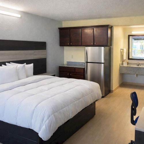 A modern hotel room with a queen bed, white bedding, dark wood furniture, a small kitchenette with a fridge, desk, and chair, and a doorway to a bathroom area.