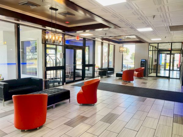 A modern hotel lobby with glass walls, bright lighting, and a mix of red curved chairs and black sofas, welcoming seating and a clean tiled floor.