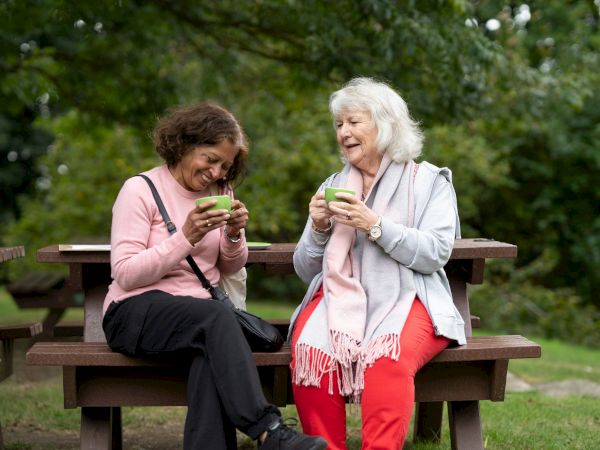 Two elderly women sit on a picnic bench outdoors, smiling and sharing drinks in a park while chatting.