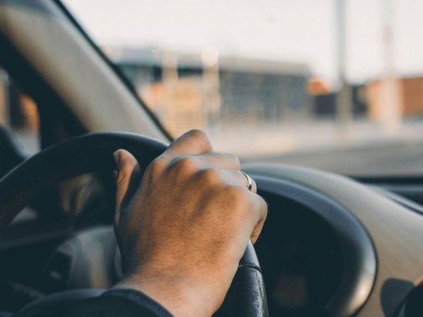 A person grips a car steering wheel inside a vehicle, with a blurred outdoor scene visible through the windshield and side window.
