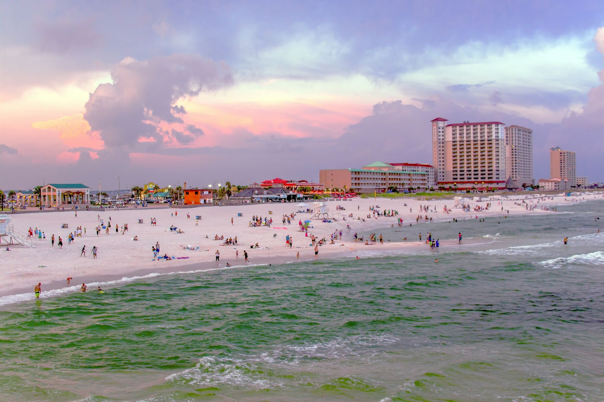 A crowded beach at sunset with people strolling along the shore, colorful buildings in the background, and gentle waves lapping the sand.