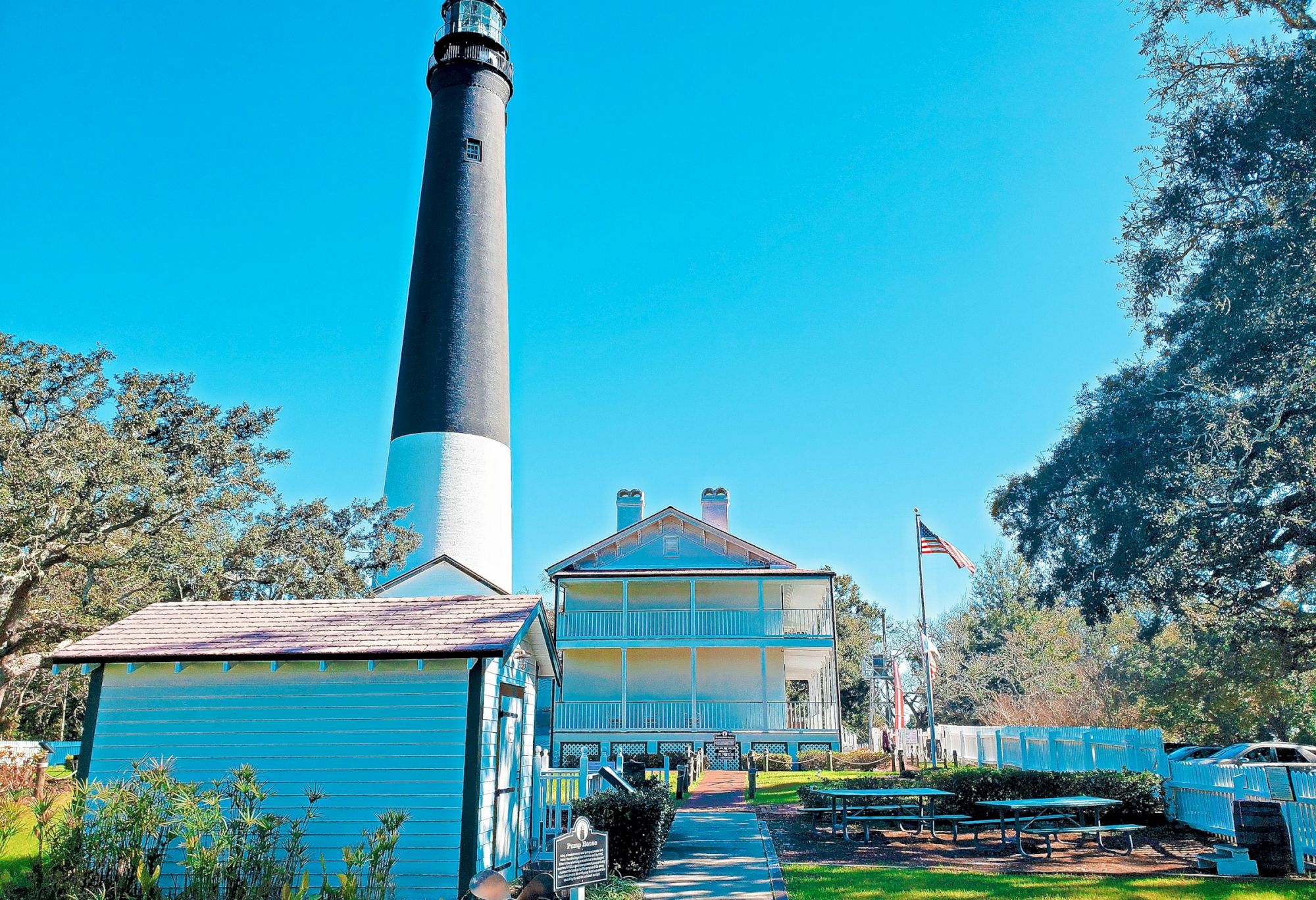 A tall black-and-white lighthouse stands behind a white house, with a small blue shed, benches, and an American flag in a sunny, leafy yard.