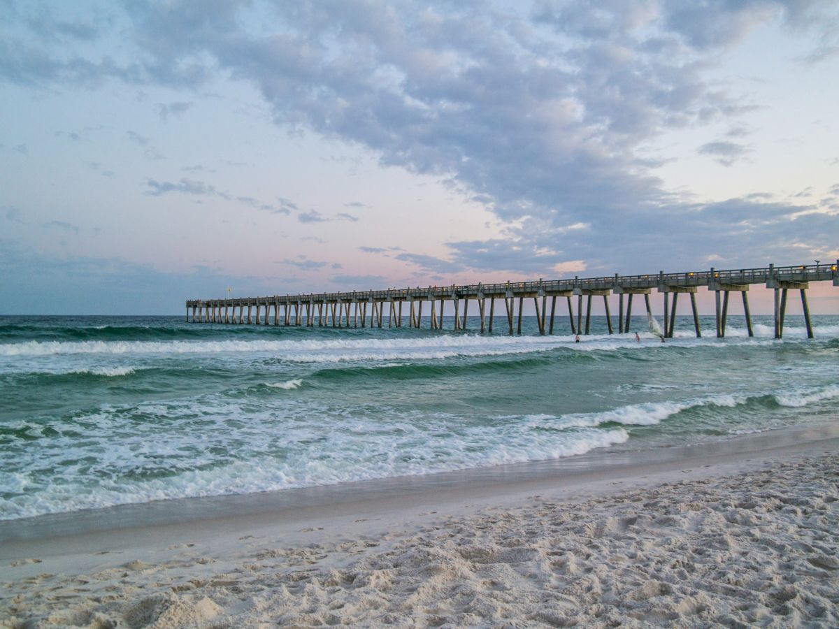A sandy beach with gentle waves and a long wooden pier extending into the ocean, under a partly cloudy sky.