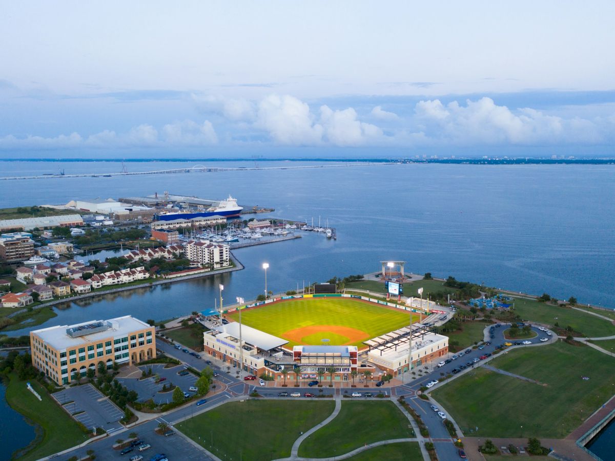An aerial view of a coastal stadium complex with bright lights, surrounding buildings, and a waterfront marina along a calm blue sea, under a cloudy sky.