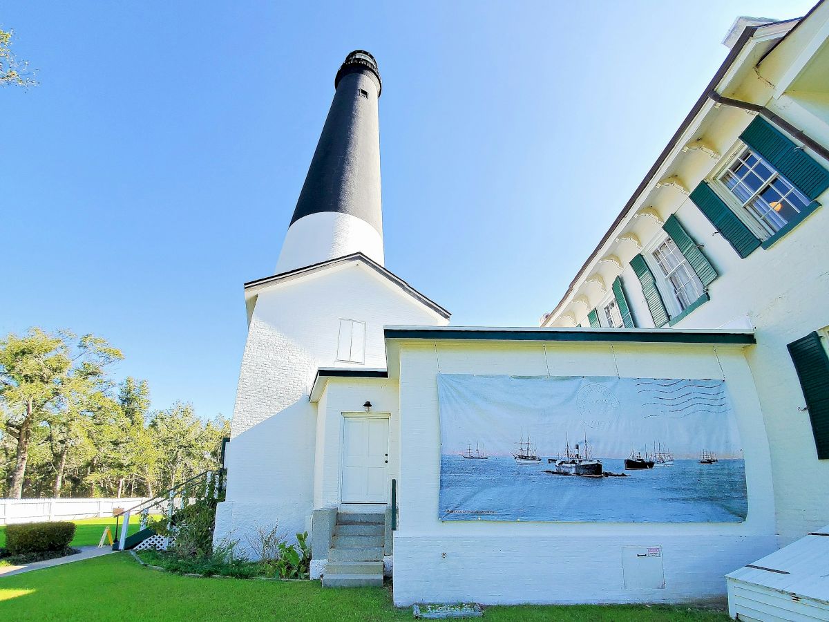 A white lighthouse with a black top stands beside a modern building under a bright blue sky, with green grass and a mural on the wall. End.