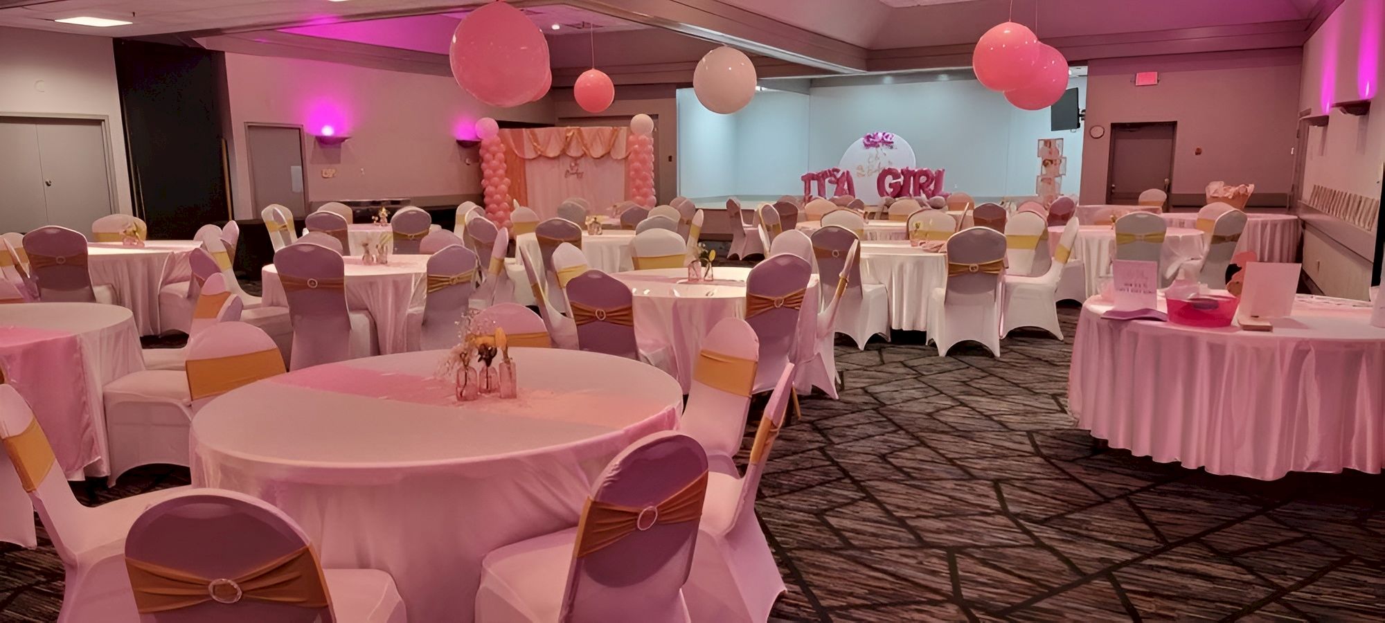 A banquet hall decorated in pink and white for a party, with round tables, white chair covers, pink sashes, balloons, and a stage at the back.