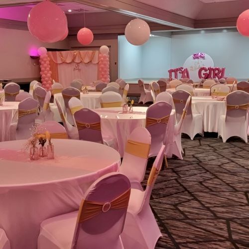 A pastel party setup: round tables with white tablecloths, pink chairs, pink and white balloons, soft pink lighting, a dessert area in the back, celebration vibe.