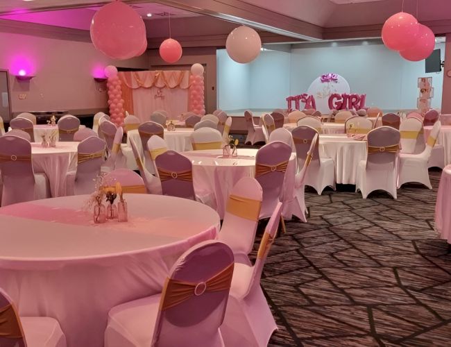 A pastel party setup: round tables with white tablecloths, pink chairs, pink and white balloons, soft pink lighting, a dessert area in the back, celebration vibe.