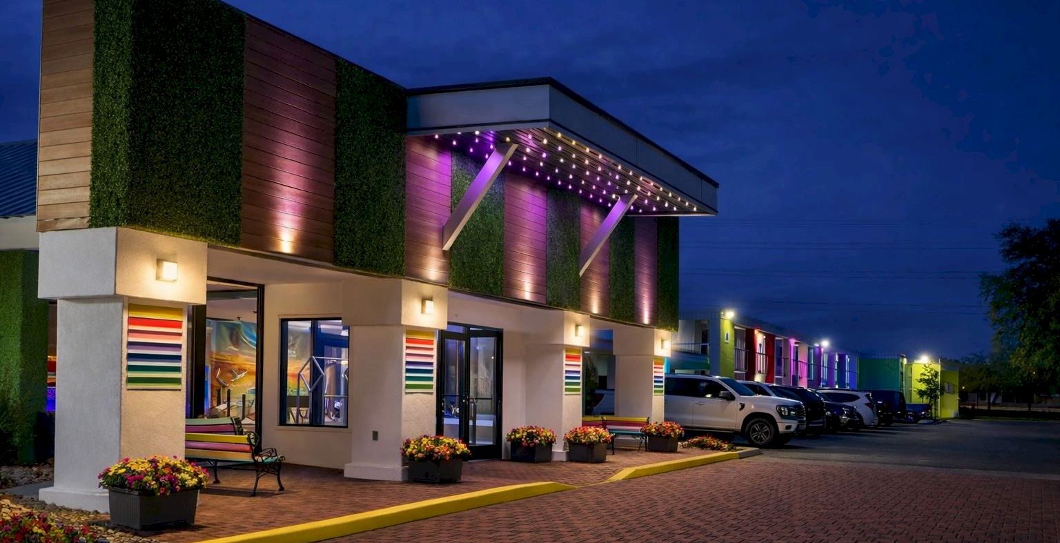 A brightly lit motel at dusk with colorful exterior lights, a row of rooms, and a parking lot along a brick-paved driveway.
