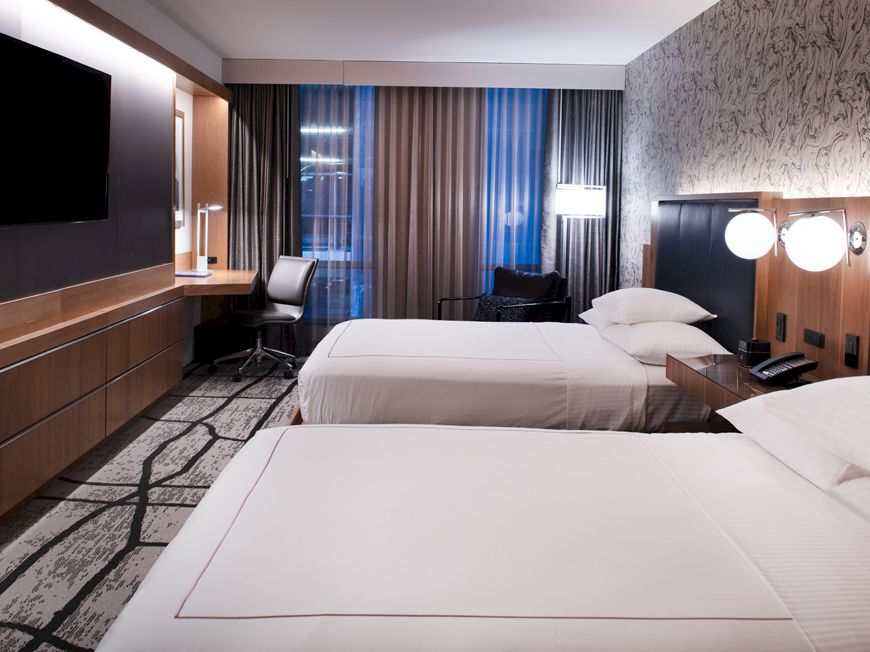 A modern hotel room with two neatly made beds, dark wood panels, and large curtains behind them.