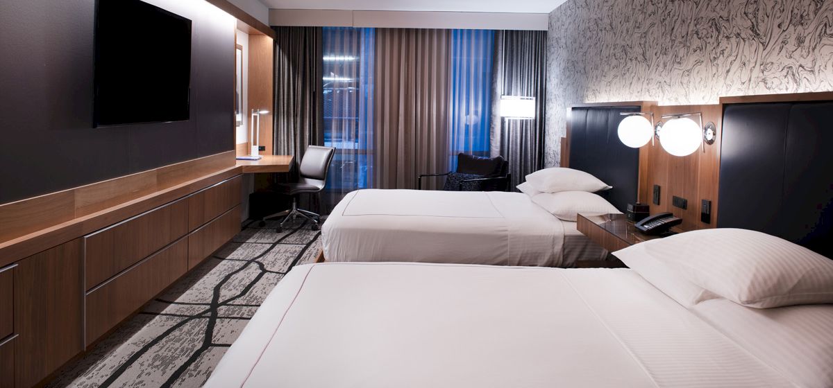 A modern hotel room with two neatly made beds, dark wood panels, and large curtains behind them.