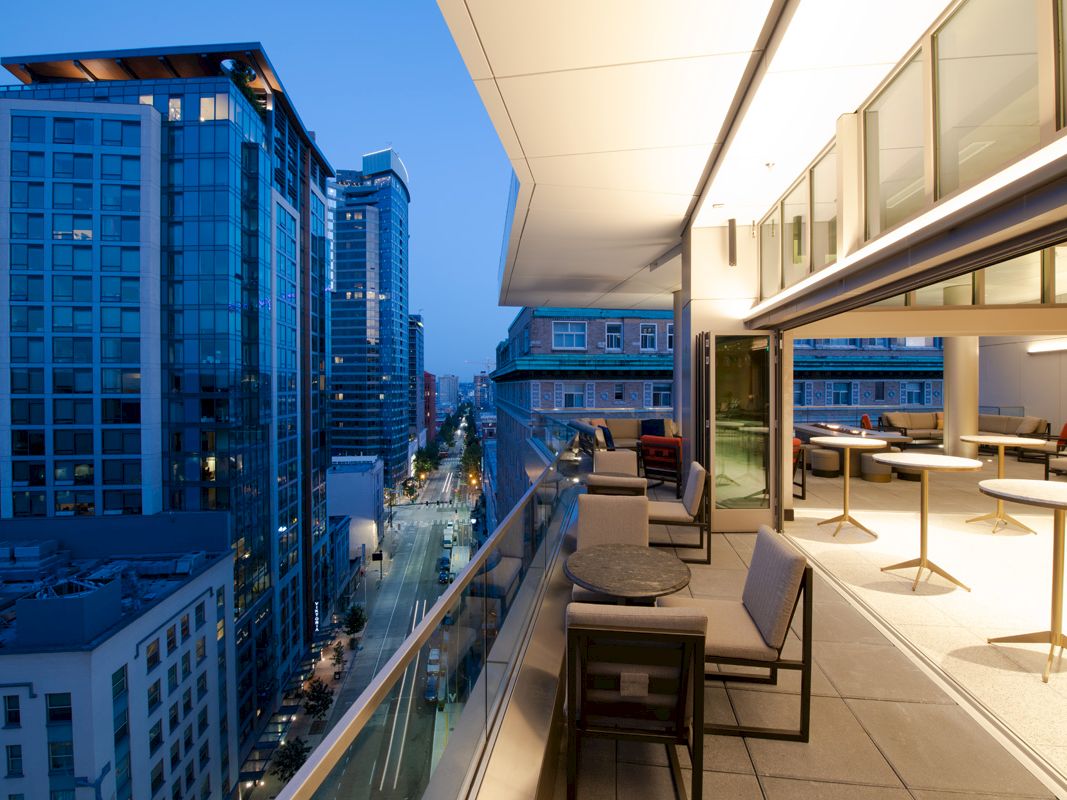 Rooftop balcony lounge with chairs and city street skyline view between tall modern buildings at dusk.