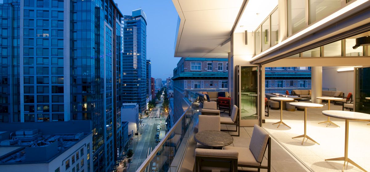 Rooftop balcony lounge with chairs and city street skyline view between tall modern buildings at dusk.